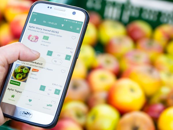 Hand hält Smartphone mit Apfelbestell-App vor einem Korb voller Äpfel