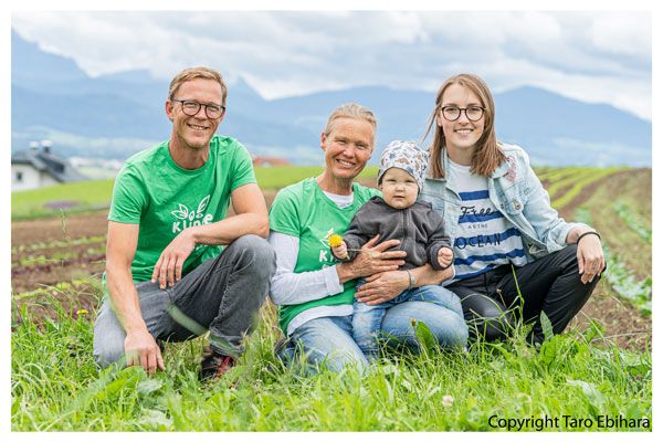 Familie mit zwei Erwachsenen, einem Kind und Baby sitzt im grünen Feld vor Bergen unter bewölktem Himmel