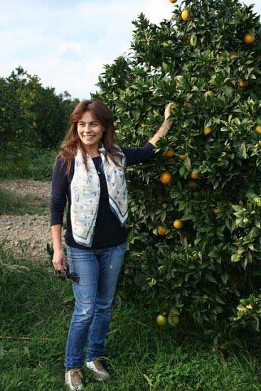 Frau steht lächelnd neben einem Orangenbaum und pflückt eine Orange im Obstgarten.
