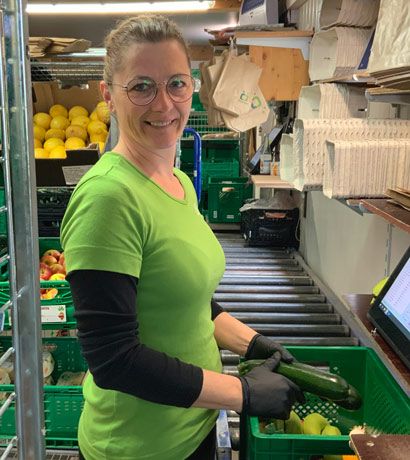 Frau in grünem T-Shirt sortiert Gemüse in Lager mit Obstkisten und Verpackungsmaterial