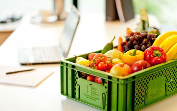 Grüne Kiste mit frischem Obst und Gemüse neben Laptop und Notizblock auf hellem Tisch