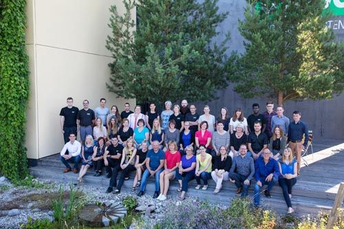 Gruppenfoto von 38 Personen vor einer Gebäudewand mit Bäumen und Pflanzen im Freien.