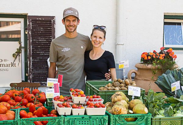 Junges Paar lächelt hinter Gemüsestand mit Tomaten, Kartoffeln und Kohl auf Bauernmarkt