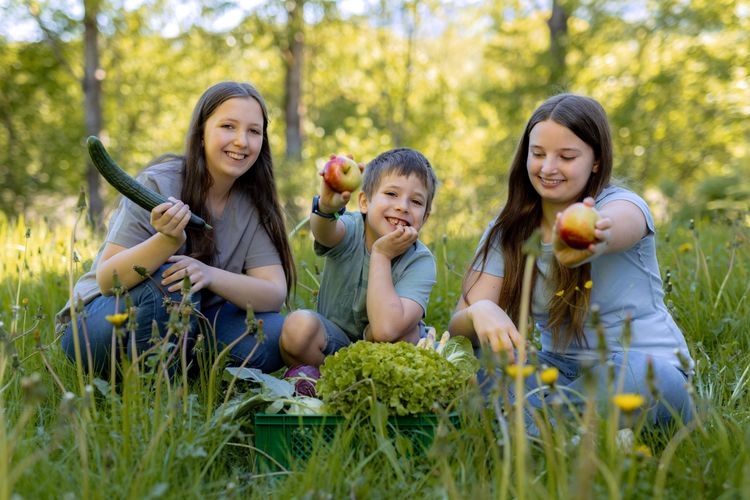 Drei lachende Kinder sitzen auf einer Wiese und zeigen Äpfel und eine Gurke mit frischem Gemüse.