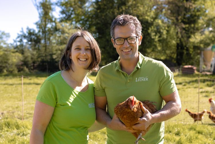 Ein lächelndes Paar im grünen T-Shirt hält ein braunes Huhn im Freien auf einer Wiese mit Hühnern im Hintergrund