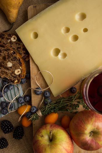 Ein großer Laib Schweizer Käse mit Löchern, umgeben von Äpfeln, Beeren, Brot und Wein.