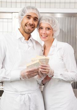 Zwei Bäcker in weißer Arbeitskleidung und Haarnetz halten frisches Brot in einer Bäckerei